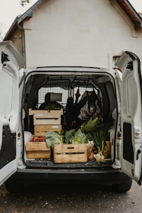 An open van parked outside a building is filled with crates of fresh vegetables, including cabbages, onions, and red bell peppers. The setup suggests an organized distribution or delivery of produce.