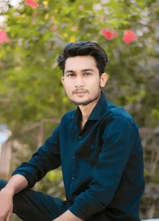 A young man with a calm expression is wearing a dark blue shirt and sitting in front of a blurred background filled with green foliage and pink flowers.