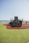 A playground structure designed to look like a ship is situated on artificial turf near a beach. The clear blue sky and a calm sea are visible in the background, with some colorful flags and poles nearby. The play area features both green and brown surface materials, likely for safety.
