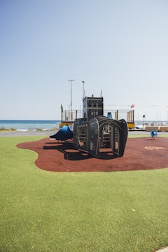 A playground structure designed to look like a ship is situated on artificial turf near a beach. The clear blue sky and a calm sea are visible in the background, with some colorful flags and poles nearby. The play area features both green and brown surface materials, likely for safety.