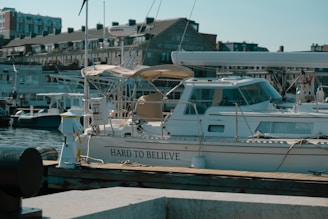 A marina with several boats docked along the water. In the foreground, a sailboat with the name 'Hard to Believe' is moored, featuring a beige canopy and a white hull. The background has a row of brick buildings with multiple floors, adding an urban element to the scene.