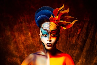 A person with elaborate body paint featuring a symmetrical design of bright orange, yellow, blue, and red colors, simulating flames and celestial elements. The makeup covers the face and upper body, with feather-like extensions on the head adding to the dramatic and artistic presentation. The background is a textured mix of dark reds and oranges, enhancing the vibrant contrast of the subject.