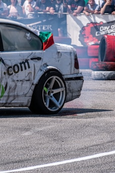 A car with visible wear and tear is performing a drift maneuver on an asphalt surface, emitting smoke from the tires. A crowd of spectators is visible in the background behind a barrier, some with banners and signs. The car has a flag draped over it, adding to the dramatic scene.