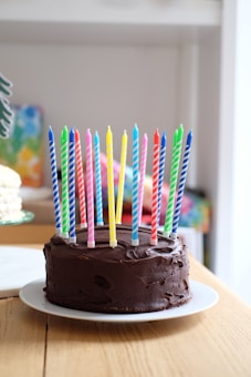 A chocolate cake covered with colorful striped candles is placed on a white plate. The cake sits on a wooden table with a blurred background featuring other colorful elements.