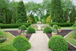 A beautifully landscaped garden featuring a walkway lined with perfectly trimmed green shrubs and planters. In the center, a large decorative fountain surrounded by vibrant flowers adds elegance. Tall trees and dense hedges create a lush backdrop, enhancing the peaceful atmosphere.
