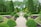 A beautifully landscaped garden featuring a walkway lined with perfectly trimmed green shrubs and planters. In the center, a large decorative fountain surrounded by vibrant flowers adds elegance. Tall trees and dense hedges create a lush backdrop, enhancing the peaceful atmosphere.