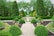A beautifully landscaped garden featuring a walkway lined with perfectly trimmed green shrubs and planters. In the center, a large decorative fountain surrounded by vibrant flowers adds elegance. Tall trees and dense hedges create a lush backdrop, enhancing the peaceful atmosphere.