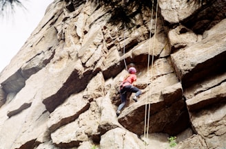 A person wearing a helmet and climbing gear ascends a rugged rock face. The climber is positioned midway on the rock wall, utilizing the cracks and ledges for support. The ropes are visible, indicating that the person is engaging in a secured rock climbing activity.