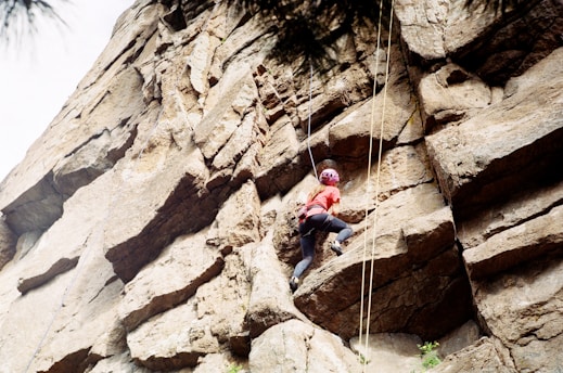A person wearing a helmet and climbing gear ascends a rugged rock face. The climber is positioned midway on the rock wall, utilizing the cracks and ledges for support. The ropes are visible, indicating that the person is engaging in a secured rock climbing activity.