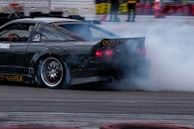 A sports car is drifting on a track, creating a large cloud of smoke from its rear tires. The vehicle is sleek and appears to be finely tuned, with visible branding on its body. The setting is an organized drift event, indicated by the presence of safety barriers and spectators in the background.