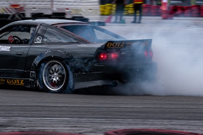 A sports car is drifting on a track, creating a large cloud of smoke from its rear tires. The vehicle is sleek and appears to be finely tuned, with visible branding on its body. The setting is an organized drift event, indicated by the presence of safety barriers and spectators in the background.