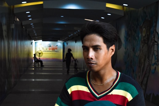 A person with a thoughtful expression stands in a dimly lit tunnel. The tunnel has decorative walls and ceiling lights leading to a bright area at the end. Silhouettes of other people can be seen, one sitting on a chair and another with a bicycle.