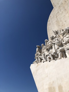 A large stone monument featuring sculptures of people in historical attire, set against a clear blue sky. The figures are intricately detailed and appear to be part of a larger architectural structure.