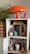 A bookshelf displays a collection of books and various decorative items. A bright orange mushroom-shaped lamp sits on top, next to small figurines of various shapes and colors. A potted plant with green foliage adds a touch of nature. The shelves contain books, DVDs, a candle, a cup, and action figures, creating a cozy and eclectic atmosphere.