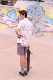 A person wearing a white short-sleeve shirt with a pattern and gray shorts is standing on a concrete surface, holding a skateboard. They are looking towards a graffiti-covered wall in the background. The graffiti features various abstract designs in bright colors including blue, yellow, and pink.