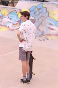 A person wearing a white short-sleeve shirt with a pattern and gray shorts is standing on a concrete surface, holding a skateboard. They are looking towards a graffiti-covered wall in the background. The graffiti features various abstract designs in bright colors including blue, yellow, and pink.