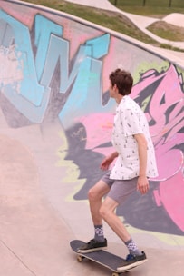 A person is skateboarding inside a graffiti-decorated skatepark. The skatepark's concrete walls are adorned with colorful graffiti featuring large, abstract letters in shades of blue, pink, and green. The skateboarder is wearing a white shirt with small patterns, gray shorts, checkered socks, and black sneakers. The environment appears open with grassy areas visible at the top edge.