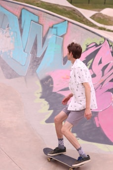 A person is skateboarding inside a graffiti-decorated skatepark. The skatepark's concrete walls are adorned with colorful graffiti featuring large, abstract letters in shades of blue, pink, and green. The skateboarder is wearing a white shirt with small patterns, gray shorts, checkered socks, and black sneakers. The environment appears open with grassy areas visible at the top edge.