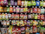 A supermarket shelf filled with various colorful packaged candies and snacks. The assortment includes well-known brands like Skittles, Haribo gummies, mints, and other assorted sweets neatly arranged. Each product has a visible price label, and the packaging features vibrant colors and eye-catching designs.