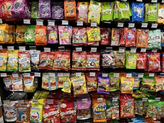 A supermarket shelf filled with various colorful packaged candies and snacks. The assortment includes well-known brands like Skittles, Haribo gummies, mints, and other assorted sweets neatly arranged. Each product has a visible price label, and the packaging features vibrant colors and eye-catching designs.