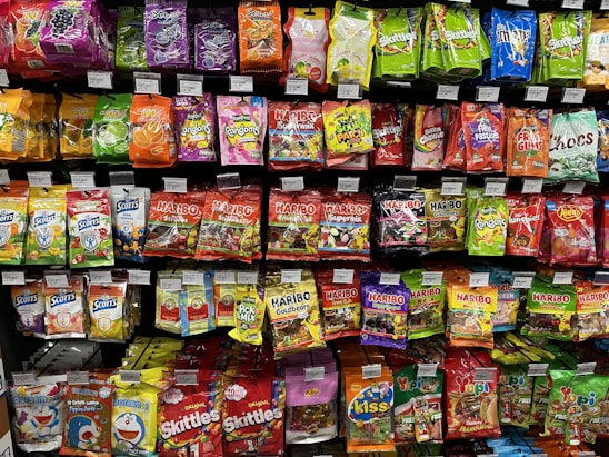 A supermarket shelf filled with various colorful packaged candies and snacks. The assortment includes well-known brands like Skittles, Haribo gummies, mints, and other assorted sweets neatly arranged. Each product has a visible price label, and the packaging features vibrant colors and eye-catching designs.