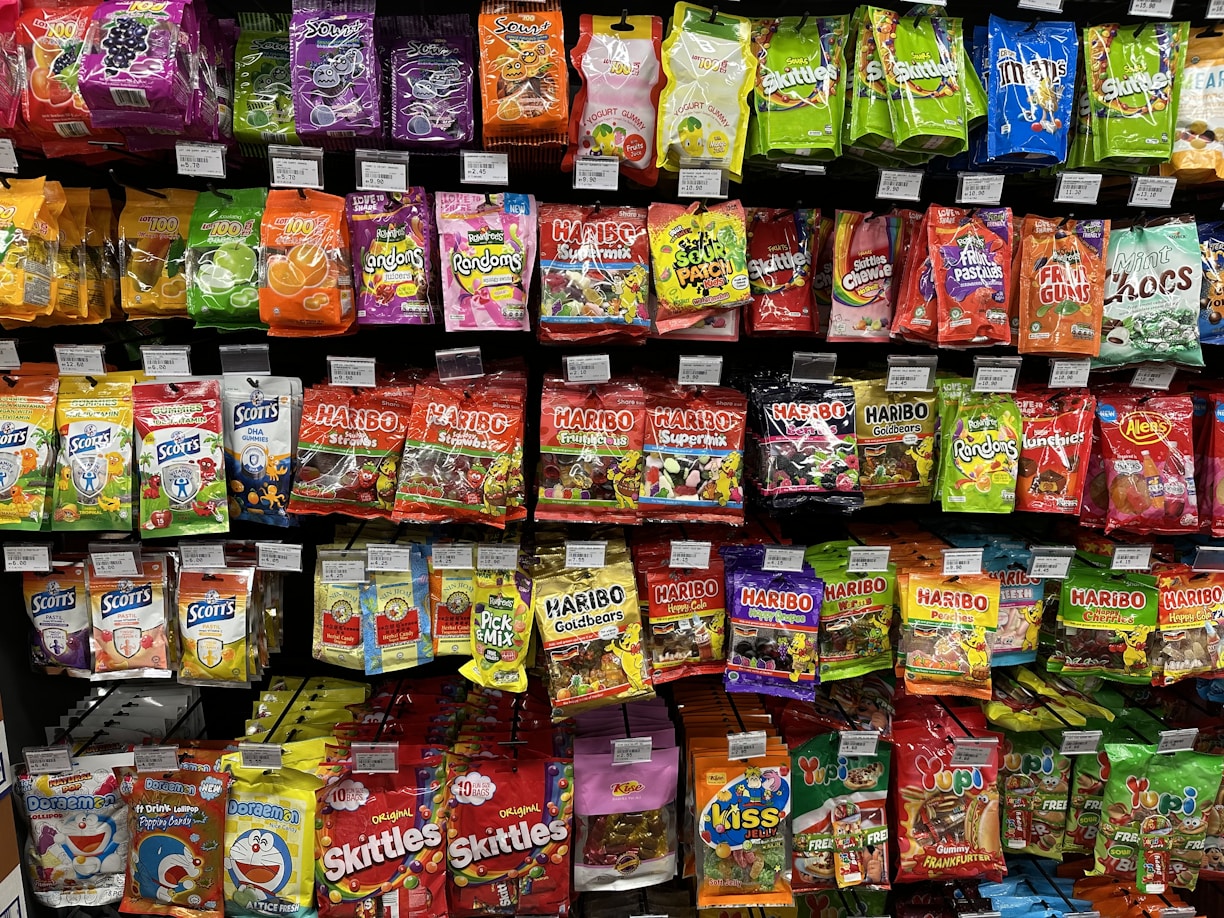 A supermarket shelf filled with various colorful packaged candies and snacks. The assortment includes well-known brands like Skittles, Haribo gummies, mints, and other assorted sweets neatly arranged. Each product has a visible price label, and the packaging features vibrant colors and eye-catching designs.