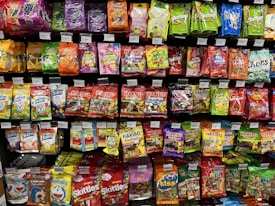 A supermarket shelf filled with various colorful packaged candies and snacks. The assortment includes well-known brands like Skittles, Haribo gummies, mints, and other assorted sweets neatly arranged. Each product has a visible price label, and the packaging features vibrant colors and eye-catching designs.