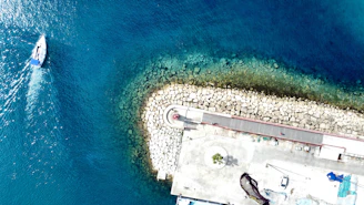 Aerial view of a marina area where a small sailboat navigates through the deep blue waters. There is a curved stone jetty with a red and white lighthouse at its center, surrounded by a circular pattern of stones. Adjacent to the lighthouse, a paved area leads to various structures, including a few parked vehicles and greenery.