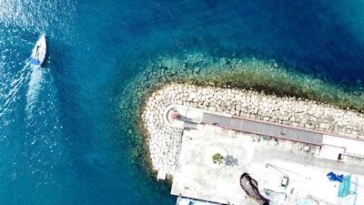 Aerial view of a marina area where a small sailboat navigates through the deep blue waters. There is a curved stone jetty with a red and white lighthouse at its center, surrounded by a circular pattern of stones. Adjacent to the lighthouse, a paved area leads to various structures, including a few parked vehicles and greenery.