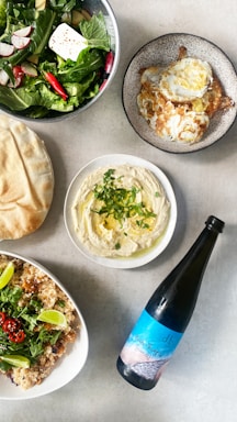 A diverse spread of dishes arranged on a table, featuring a fresh green salad with radishes, cheese, and a chili pepper, a plate of fried eggs, a bowl of hummus garnished with herbs and olive oil, flatbread on the side, a dish of couscous with greens and lime wedges, and a bottle of wine.
