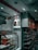 An interior shot of a retail store features shelves stocked with various products. A glass display case showcases red-packaged items. Overhead, promotional signs advertise cold beverages like coffee and Frappe Mocha. The store's aesthetic includes white tiled walls, and red accents mark the space with non-vegetarian options highlighted in red signage.