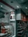 An interior shot of a retail store features shelves stocked with various products. A glass display case showcases red-packaged items. Overhead, promotional signs advertise cold beverages like coffee and Frappe Mocha. The store's aesthetic includes white tiled walls, and red accents mark the space with non-vegetarian options highlighted in red signage.