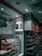 An interior shot of a retail store features shelves stocked with various products. A glass display case showcases red-packaged items. Overhead, promotional signs advertise cold beverages like coffee and Frappe Mocha. The store's aesthetic includes white tiled walls, and red accents mark the space with non-vegetarian options highlighted in red signage.