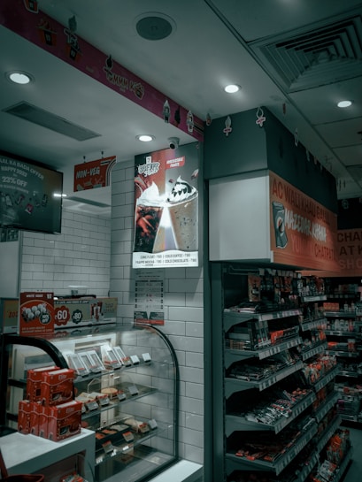 An interior shot of a retail store features shelves stocked with various products. A glass display case showcases red-packaged items. Overhead, promotional signs advertise cold beverages like coffee and Frappe Mocha. The store's aesthetic includes white tiled walls, and red accents mark the space with non-vegetarian options highlighted in red signage.