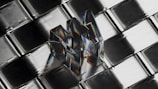 A transparent, reflective object shaped like the letter 'M' is placed on a metallic, grid-like surface. The object reflects a spectrum of colors, creating a prismatic effect, and appears three-dimensional against the geometric pattern of the background.