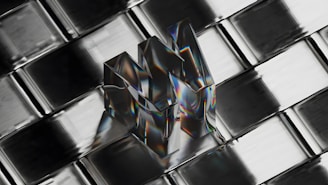 A transparent, reflective object shaped like the letter 'M' is placed on a metallic, grid-like surface. The object reflects a spectrum of colors, creating a prismatic effect, and appears three-dimensional against the geometric pattern of the background.