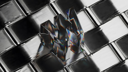 A transparent, reflective object shaped like the letter 'M' is placed on a metallic, grid-like surface. The object reflects a spectrum of colors, creating a prismatic effect, and appears three-dimensional against the geometric pattern of the background.