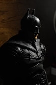 A figure of a person dressed in a black armored suit with a mask and pointed ears, resembling a superhero. The lighting creates shadows in the background, providing a dark and moody atmosphere.