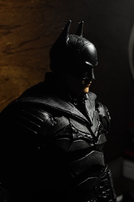 A figure of a person dressed in a black armored suit with a mask and pointed ears, resembling a superhero. The lighting creates shadows in the background, providing a dark and moody atmosphere.