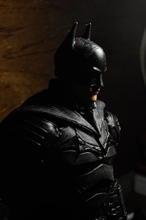 A figure of a person dressed in a black armored suit with a mask and pointed ears, resembling a superhero. The lighting creates shadows in the background, providing a dark and moody atmosphere.