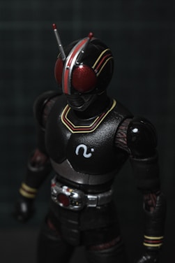 A detailed action figure resembling a robotic superhero with large red eyes, a black helmet with red and yellow striping, and a suit with mechanical details. The figure has antennas protruding from its head and a prominent belt with intricate designs.