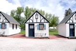 Three quaint, small cottages with a charming, old-fashioned design featuring white stucco walls and black timber trims. Each cottage has a blue door and window frames adorned with flower boxes. The setting includes a patio area with metal chairs and tables under a partly cloudy sky, surrounded by lush green trees.