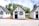 Three quaint, small cottages with a charming, old-fashioned design featuring white stucco walls and black timber trims. Each cottage has a blue door and window frames adorned with flower boxes. The setting includes a patio area with metal chairs and tables under a partly cloudy sky, surrounded by lush green trees.