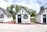 Three quaint, small cottages with a charming, old-fashioned design featuring white stucco walls and black timber trims. Each cottage has a blue door and window frames adorned with flower boxes. The setting includes a patio area with metal chairs and tables under a partly cloudy sky, surrounded by lush green trees.