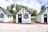 Three quaint, small cottages with a charming, old-fashioned design featuring white stucco walls and black timber trims. Each cottage has a blue door and window frames adorned with flower boxes. The setting includes a patio area with metal chairs and tables under a partly cloudy sky, surrounded by lush green trees.