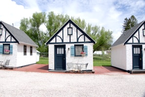Three quaint, small cottages with a charming, old-fashioned design featuring white stucco walls and black timber trims. Each cottage has a blue door and window frames adorned with flower boxes. The setting includes a patio area with metal chairs and tables under a partly cloudy sky, surrounded by lush green trees.