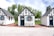 Three quaint, small cottages with a charming, old-fashioned design featuring white stucco walls and black timber trims. Each cottage has a blue door and window frames adorned with flower boxes. The setting includes a patio area with metal chairs and tables under a partly cloudy sky, surrounded by lush green trees.