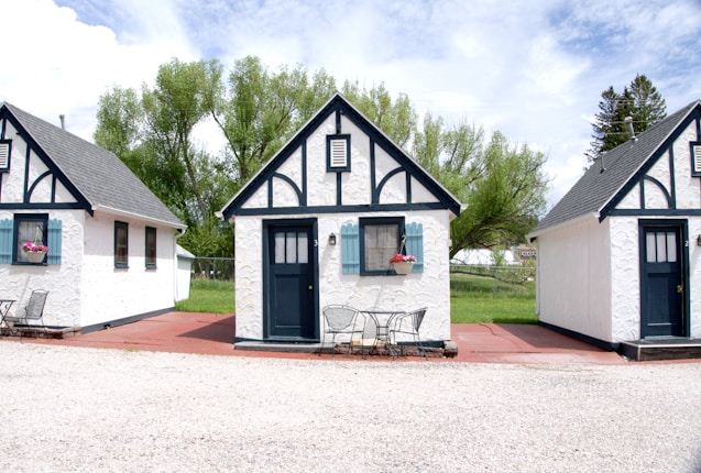 Three quaint, small cottages with a charming, old-fashioned design featuring white stucco walls and black timber trims. Each cottage has a blue door and window frames adorned with flower boxes. The setting includes a patio area with metal chairs and tables under a partly cloudy sky, surrounded by lush green trees.
