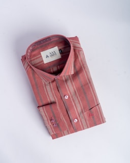 A neatly folded, striped shirt in shades of red and beige, featuring a tag with branding inside the collar. The shirt has a classic design, including buttons and a pocket detail on a smooth, neutral background.