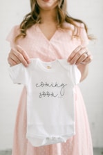 A person is holding a small white baby onesie with the words 'coming soon...' printed on it. The person appears to be in soft focus, wearing a light pink dress with a subtle pattern. The background is neutral, enhancing the focus on the onesie.